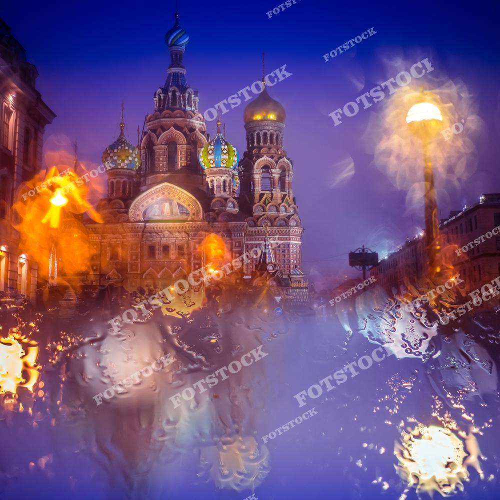 На фотографии изображен храм Спаса-на-Крови в Санкт-Петербурге, видимый через стекло, покрытое дождевыми каплями. Вечерний свет добавляет яркие блики и отражения, создавая эффект таинственности и уюта. Спас-на-Крови — одно из самых узнаваемых зданий города, построенное в традиционном русском стиле с яркими куполами. Этот храм расположен на канале Грибоедова и привлекает туристов своим архитектурным великолепием.